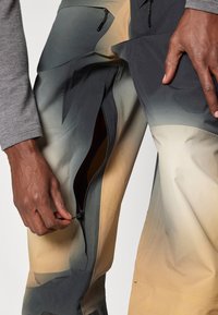 Άτομο που ξετυλίγει το πλαϊνό φερμουάρ σε παντελόνι με κλίση από μπεζ σε γκρι, ενώ είναι ορατό ένα γκρι μακρυμάνικο μπλουζάκι στο χέρι.