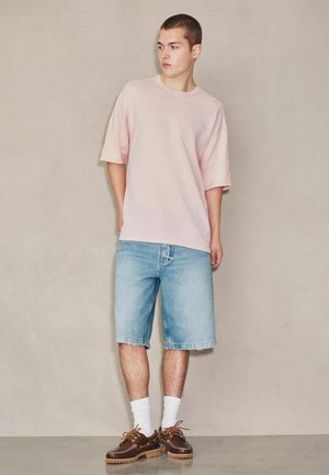 Fiatal férfi áll egy bézs fal előtt, világos rózsaszín, túlméretezett pólót, világoskék farmer rövidnadrágot, fehér zoknit és barna bőr cipőt viselve.