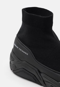 Svart stickad ankelhög sneaker med ribbad manschett, gummisole och "ARMANI EXCHANGE"-logotyp i vitt på sidan. Slät textur, stilren design.