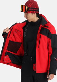 Rote und schwarze Skijacke mit einer Reißverschlusstasche, Netzfutter und verstellbaren Bündchen. Enthält Logo-Details und eine Innentasche.