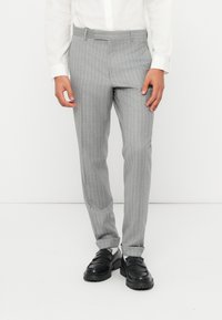 Pantaloni grigi a righe sottili, realizzati in tessuto morbido, con vestibilità sartoriale, passanti per cintura e una consistenza liscia. Abbinati a scarpe nere senza lacci.