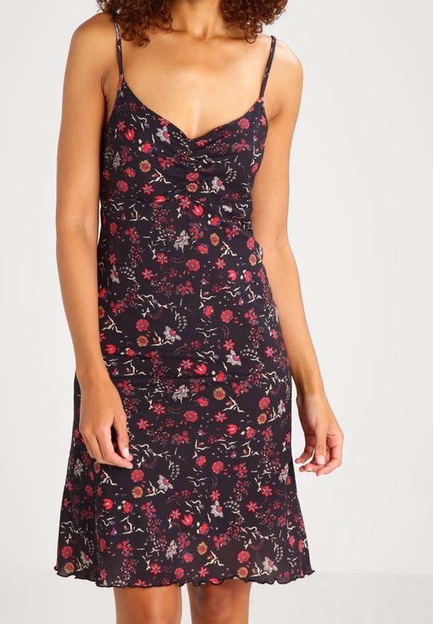 Femme portant une robe noire sans manches à fleurs avec fines bretelles et un décolleté froncé, debout contre un fond clair uni.