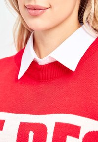 Rode trui met een geribbelde halslijn, voorzien van een wit grafisch patroon. Een witte blouse is onder de trui gelaagd.