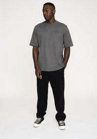 Grå bomulds t-shirt med korte ærmer og rund hals, med et lille sort logo på brystet. Parret med sorte bukser og sneakers.