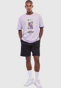 Lilla kortærmet t-shirt med grafisk print, stylede med sorte shorts. Har rund halsudskæring og afslappet pasform. Hvide sneakers fuldender outfittet.