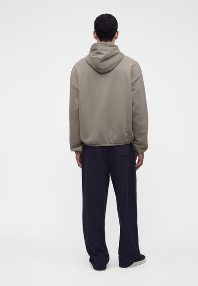 Personne portant un sweat à capuche beige ample et un pantalon large bleu marine foncé, debout de dos devant un fond clair uni.