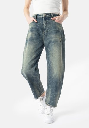 Person iført løstsiddende, falmede blå jeans, hvid tanktop og hvide sneakers, med hænderne i lommerne og det ene ben krydset over det andet.