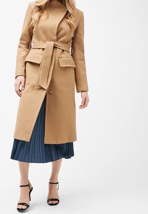Femme portant un manteau ceinturé couleur camel sur une jupe plissée bleue, assorti de sandales à talons hauts noires à brides, debout sur un fond blanc.