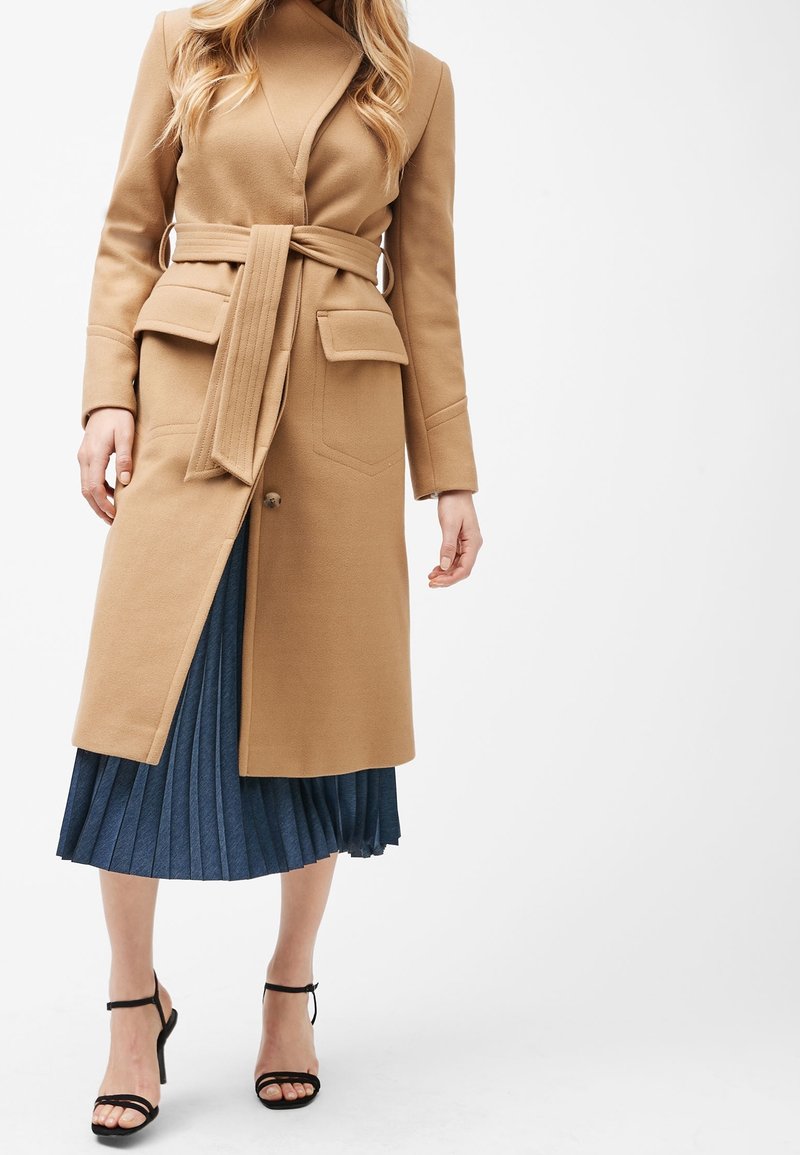 Femme portant un manteau ceinturé couleur camel sur une jupe plissée bleue, assorti de sandales à talons hauts noires à brides, debout sur un fond blanc.