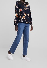 Blusa floreale blu navy con maniche lunghe e scollo a legaccio, abbinata a jeans blu a gamba dritta e sneakers bianche.