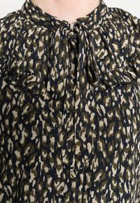 Blusa escura com padrão de leopardo em tons de olive e bege. Apresenta um colarinho com babados, um laço no decote e um tecido texturizado.