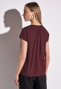 Blusa bordeaux a maniche corte con dettaglio a pieghe sul retro, in tessuto morbido, colletto rotondo e vestibilità rilassata, abbinata a pantaloni neri.