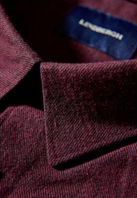 Bordeaux gekleed stof met een visgraatpatroon, voorzien van een puntige kraag en een genaaid label in marineblauw. Zacht, warm materiaal.
