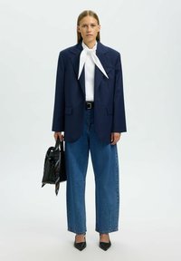 Blazer azul marino con rayas, blusa blanca con lazo, vaqueros de pierna ancha azules, tacones negros y bolso negro con pañuelo estampado.