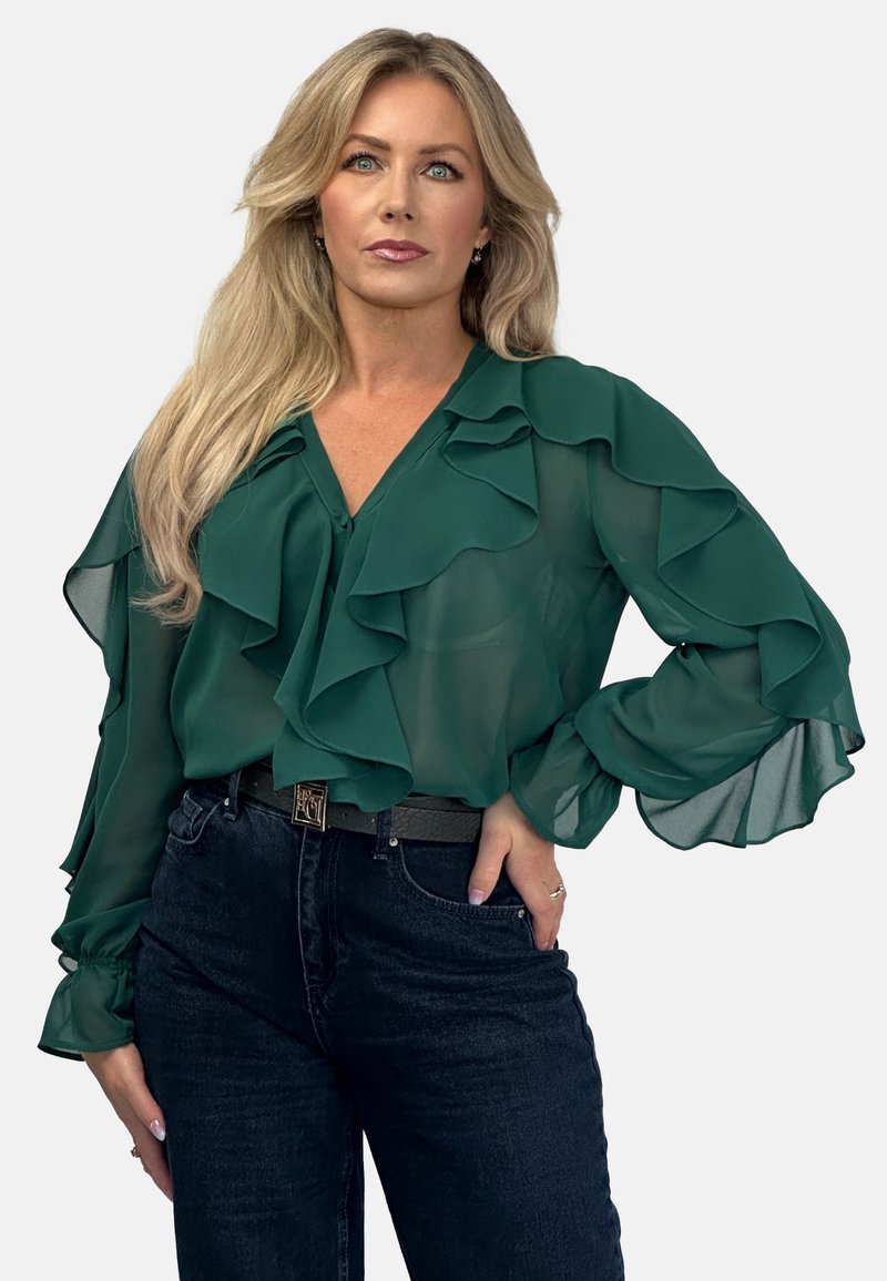 Green, sheer blouse with layered ruffles, front button closure, and long sleeves. Paired with dark denim jeans and a belt featuring a metallic accent.