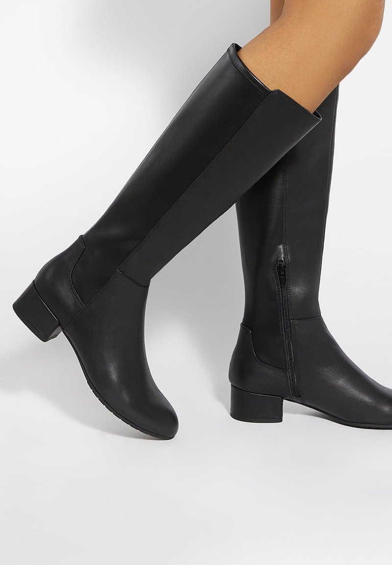 Bottes en cuir noires montantes jusqu'aux genoux avec talons blocs bas et fermetures éclair latérales, portées sur des jambes nues sur un fond clair uni.