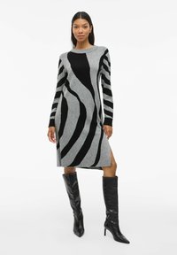 Abito maglione grigio con strisce astratte nere, maniche lunghe, scollo rotondo, spacco laterale, abbinato a stivali neri con tacco al ginocchio.
