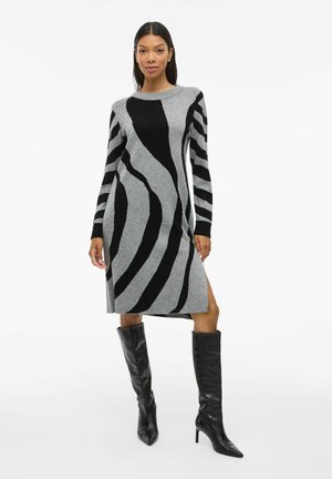 Robe pull grise avec des rayures abstraites noires, manches longues, col rond, fente sur le côté, associée à des bottes à talons noires jusqu'aux genoux.