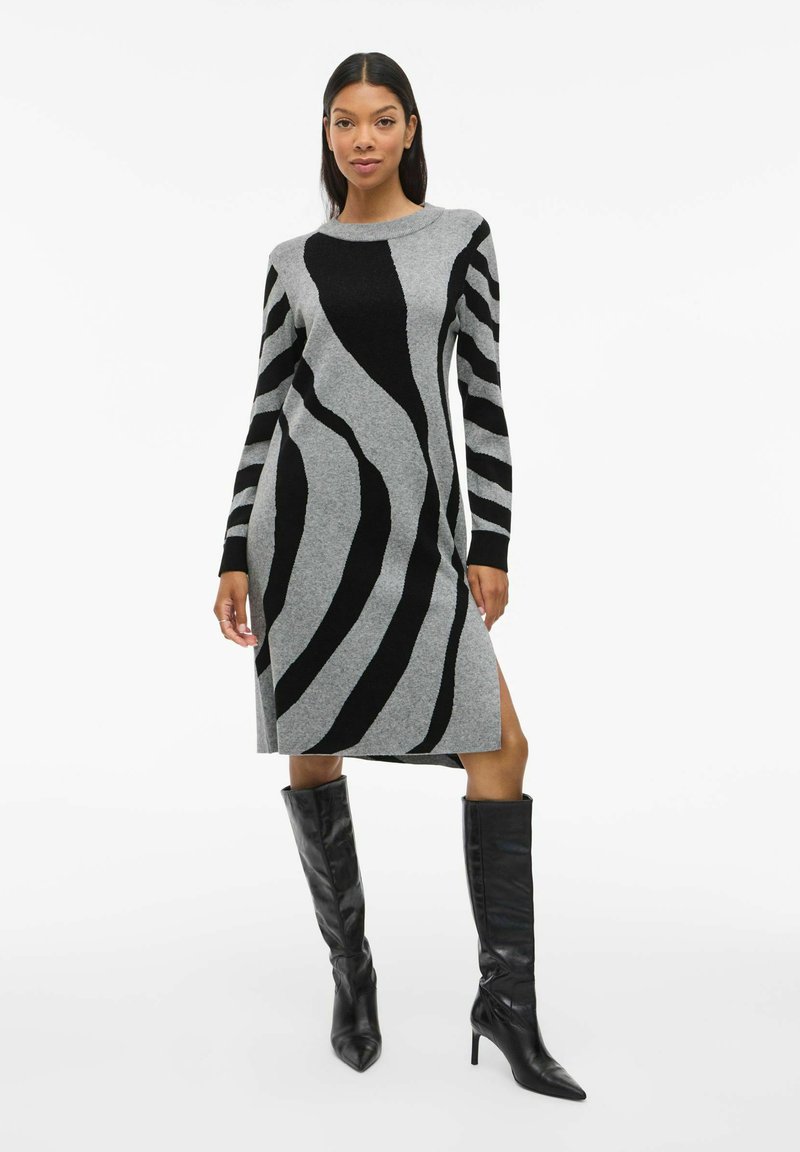 Abito maglione grigio con strisce astratte nere, maniche lunghe, scollo rotondo, spacco laterale, abbinato a stivali neri con tacco al ginocchio.