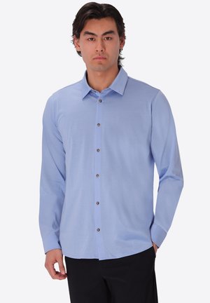 Uomo con camicia azzurro chiaro a maniche lunghe con bottoni e pantaloni neri, in piedi su uno sfondo bianco semplice.