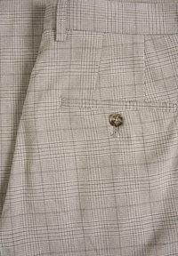 Beige ruuduline kangas, millel on tekstuuriga mustriga, tagaküljel on tasku ringikujulise nööbiga detailiga. Nähtavad õmblused.