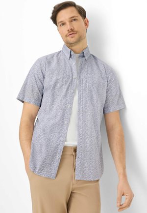 Mann trägt ein kurzärmeliges Hemd mit geometrischem Muster über einem weißen T-Shirt und beigen Hosen, steht vor einem weißen Hintergrund.