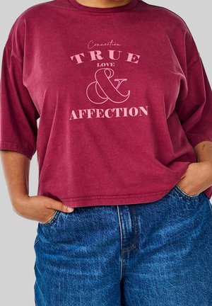 Osoba w bordowej koszuli z napisem „Connection True Love & Affection” i niebieskich dżinsach, ręce spoczywają w kieszeniach.