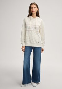 Sweatshirt à capuche crème avec un logo sur le devant, texture en forme de diamant et poche kangourou, associé à un jean bleu à jambes larges et des baskets blanches.
