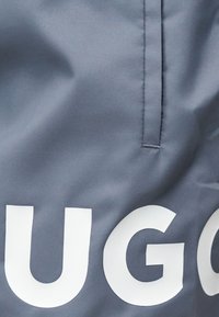 Κοντινή λήψη γκρίζου υφάσματος με μεγάλα λευκά γράμματα "UG" και κάθετη ραφή πάνω από τα γράμματα.