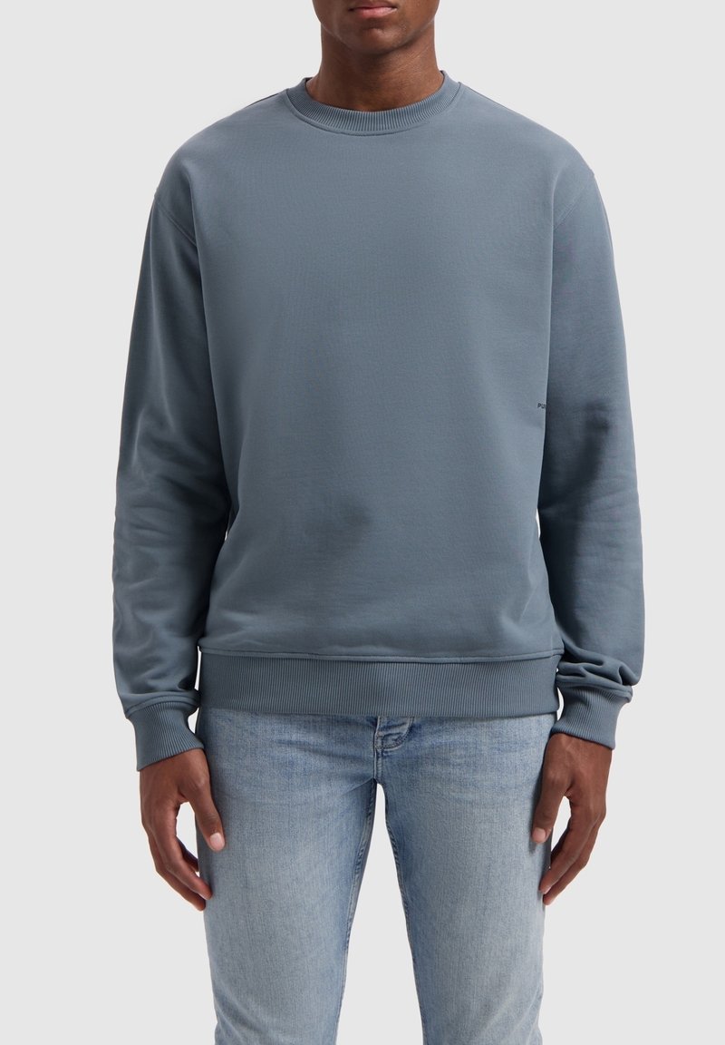 Sweat-shirt en coton gris avec col rond, poignets et ceinture côtelés. Texture lisse et design minimaliste avec un logo sur le côté.