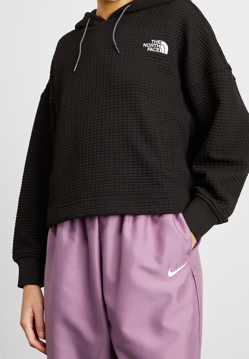 Hoodie corto nero con un design testurizzato, caratterizzato da un logo bianco sul petto. Abbinato a pantaloni larghi di un leggero colore viola.