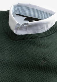 Pulover tricotat de culoare verde închis cu decolteu în V, având o cămașă albă cu guler dedesubt. Accente cu guler în dungi albastre și albe. Textură moale.