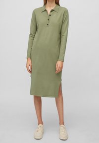 Robe polo olive à manches longues avec une patte de boutonnage, fentes latérales et une texture lisse. Associée à des baskets épaisses de couleur claire.