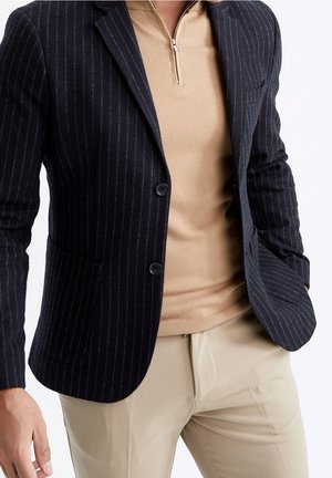 Mies pukeutunut laivastonsiniseen pinstriped bleiseriin beigen vetoketjuneuleen ja vaaleiden beigejen housujen päällä, seisoo yhdellä kädellä taskussa.