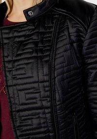 Schwarze gesteppte Jacke mit hohem Kragen, ausgestattet mit einem dezenten geometrischen Muster, Reißverschluss und einem glatten, kontrastierenden Material an den Schultern.