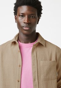 Camicia beige con bottoni e superficie testurizzata, taschino sul petto e una t-shirt girocollo rosa sotto. Design semplice, vestibilità casual.