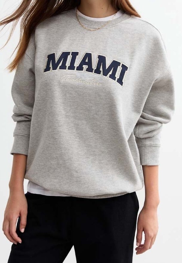 Grijze sweatshirt met de tekst "MIAMI" in grote marineblauwe letters en een klein geborduurd detail eronder. Gemaakt van zacht materiaal met geribbelde boorden.