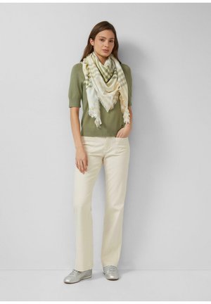 Femme debout contre un mur, portant un haut vert à manches courtes, un pantalon crème, des chaussures argentées et une grande écharpe à carreaux crème et verte avec des bords frangés.