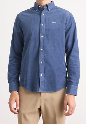 Homme portant une chemise bleue à manches longues boutonnée et un pantalon beige, debout devant un fond clair et uni.