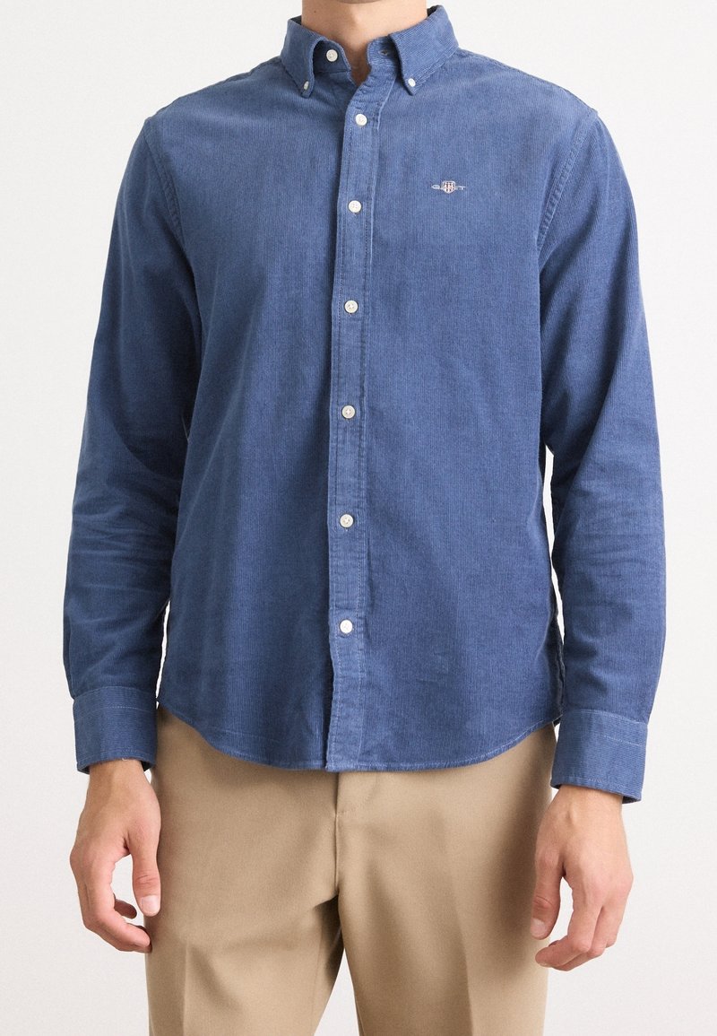 Homme portant une chemise bleue à manches longues boutonnée et un pantalon beige, debout devant un fond clair et uni.