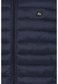 Marineblaue gesteppte Jacke vorne mit vertikalem schwarzem Reißverschluss und kleinem quadratischem Logo-Patch auf der linken Brustseite.