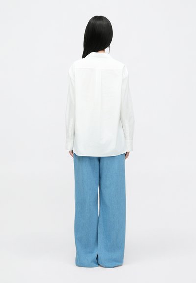 Camisa branca de botão com um comprimento maior na parte de trás, combinada com calças de ganga largas de tom azul claro, exibida de costas sobre um fundo claro.