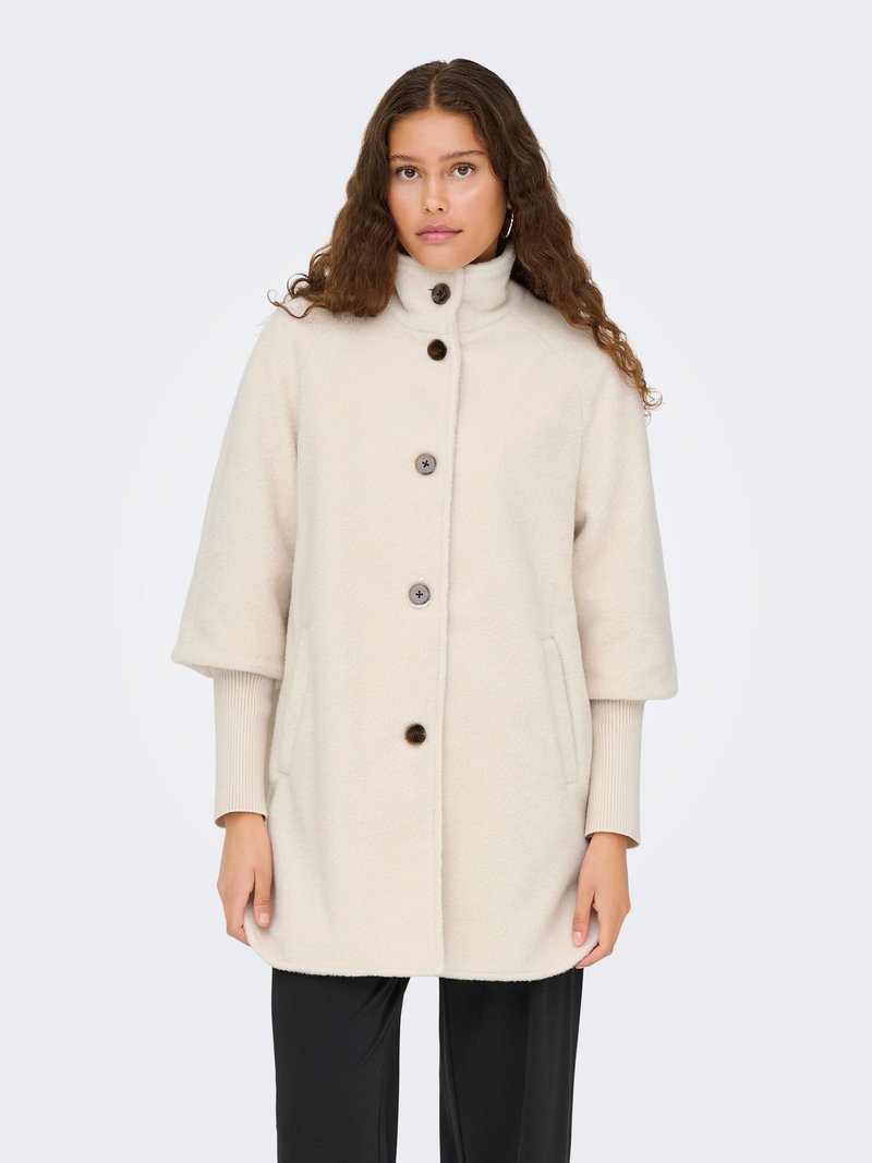 Femme portant un manteau en laine couleur crème à manches trois-quarts par-dessus un haut à manches longues et un pantalon noir, debout devant un fond uni.