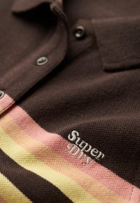 Detailní záběr na hnědý pletený polo shirt s růžovými, broskvovými a žlutými pruhy a výšivkou "Super DIY" v bílé barvě blízko oblasti hrudníku.