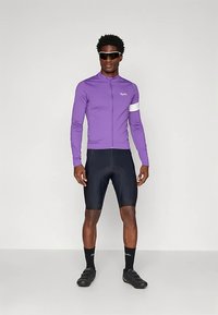 Un homme portant un maillot de cyclisme à manches longues violet, un short de cyclisme noir, des chaussettes noires, des chaussures de cyclisme noires et des lunettes de soleil noires, se tenant droit.