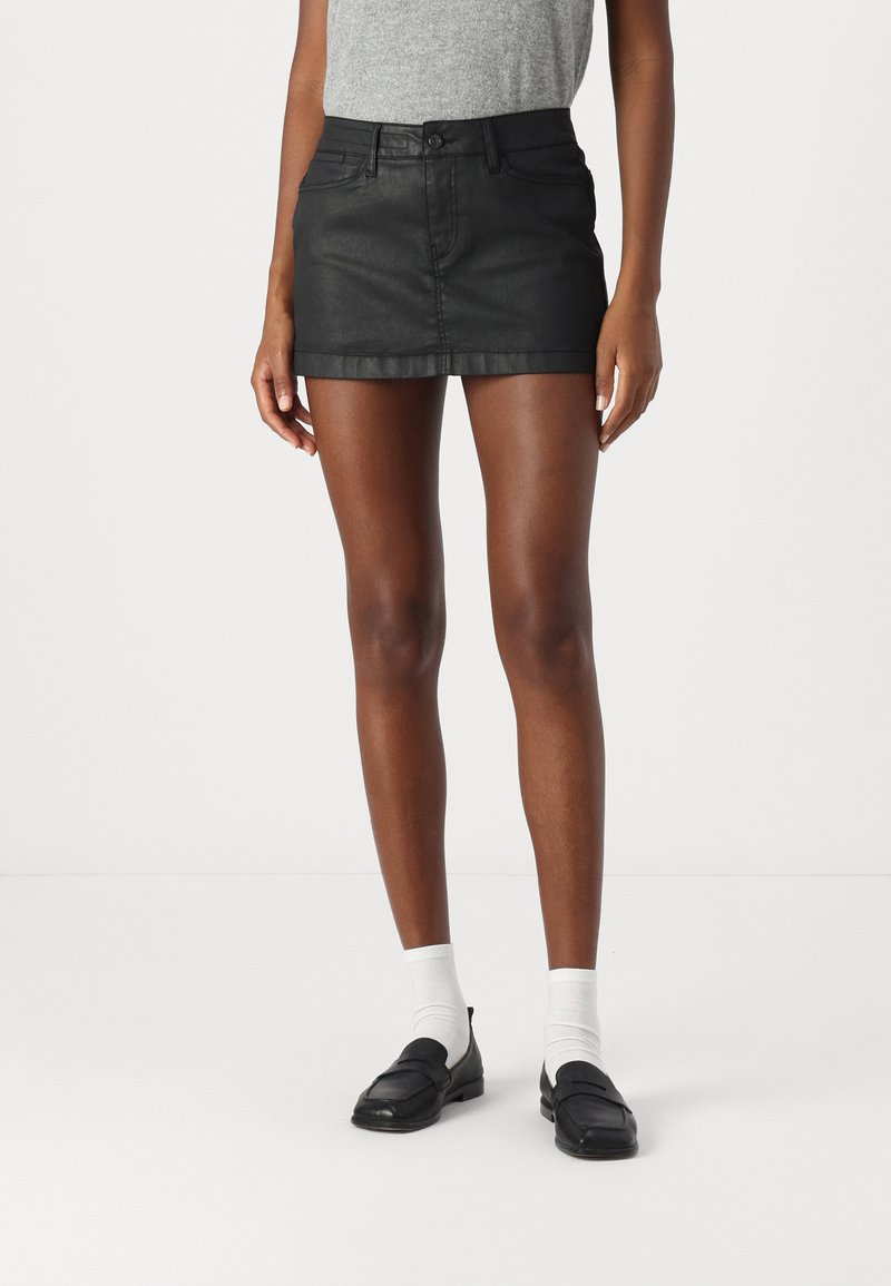 Mini jupe en denim noir avec une coupe ajustée, dotée d'une fermeture à bouton sur le devant, d'un design à cinq poches et d'un ourlet propre. Portée avec des chaussettes blanches et des mocassins.