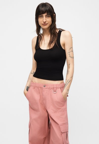 Žena s dlhými tmavými vlasmi a tetovaniami, ktorá má na sebe čierny tielkový crop top a voľné ružové cargo nohavice, ruky vreckách.
