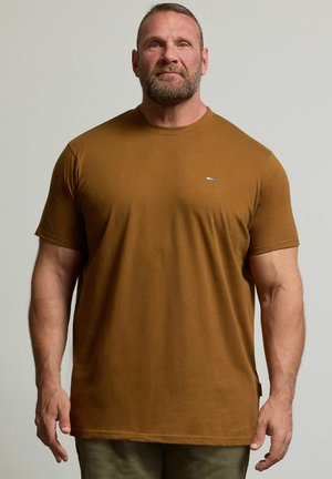 Baardige man met kort haar draagt een aansluitend bruin T-shirt met korte mouwen en staat tegen een effen lichte achtergrond.