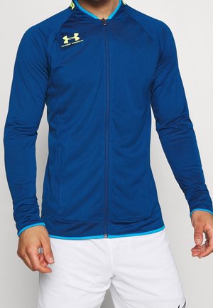 Mann trägt eine blaue Under Armour Jacke mit Reißverschluss und neongelbem Logo, kombiniert mit weißen Shorts vor einfachem Hintergrund.