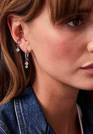 Silverfärgade hoopsörhängen med strukturerad yta, prydda med små klara ädelstenar som hänger från den nedre delen. Bärs på en ljus hudton.